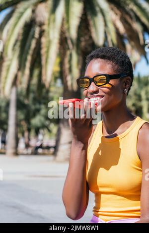 elegante donna africana che invia messaggi vocali all'aperto in una giornata di sole. Indossa un abbigliamento casual estivo e sorride felicemente Foto Stock