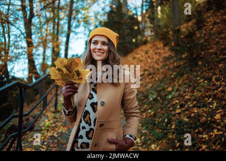 Ciao autunno. Giovane donna sorridente in cappotto beige e cappello arancione con foglie di giallo autunno fuori sul parco della città in autunno. Foto Stock