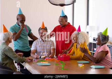 Allegro multirazziale seniors indossare cappelli partito mentre festeggia il compleanno dell'amico in casa di cura Foto Stock