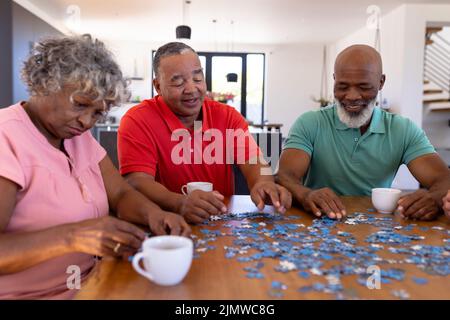 Amici senior multirazziale che organizzano pezzi di puzzle sul tavolo mentre si gode il caffè in casa di cura Foto Stock