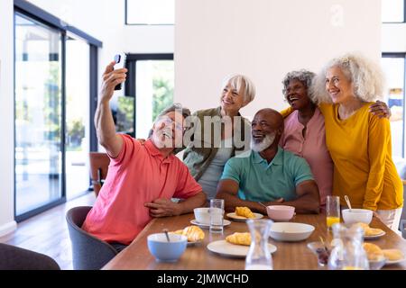L'anziano caucasico prende selfie con amici multirazziali mentre fa colazione al tavolo da pranzo Foto Stock