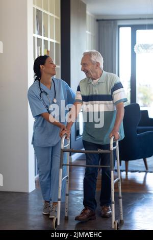 Fisioterapista femminile biracale che parla e aiuta l'anziano caucasico a camminare con il camminatore Foto Stock