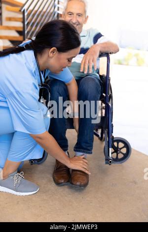 Fisioterapista femminile biracale che regola le gambe dell'anziano caucasico seduto su sedia a rotelle a casa Foto Stock