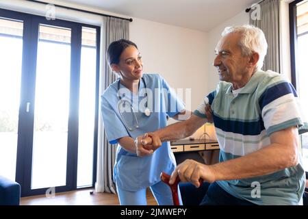 Fisioterapista femminile biracale che assiste l'uomo maggiore caucasico in piedi con la canna da camminare a casa Foto Stock