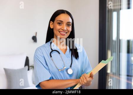 Ritratto di fisioterapista femminile felice biraciale con rapporto e stetoscopio in piedi a casa Foto Stock
