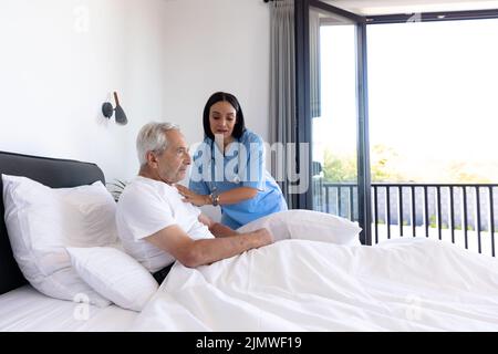 Fisioterapista femminile biracale che aiuta l'uomo maggiore caucasico a sdraiarsi a letto a casa, spazio copia Foto Stock