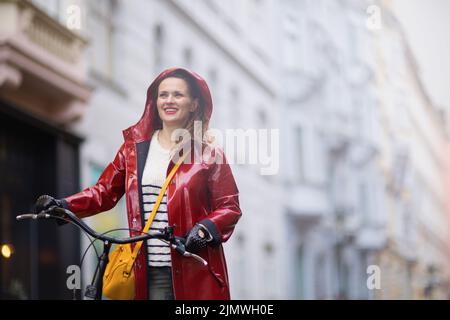 felice donna elegante in cappotto rosso con bicicletta sotto la pioggia fuori in città. Foto Stock