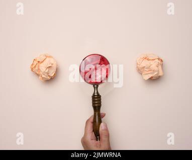 La mano femminile tiene una lente d'ingrandimento in legno e palle di carta stropicciate su sfondo beige Foto Stock