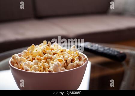 Ciotola con popcorn e telecomando tv su tavolo in vetro vicino al divano Foto Stock