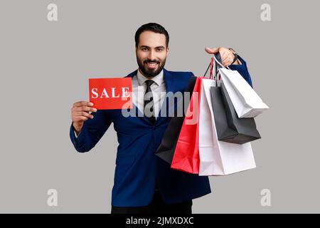 Uomo bearded sorridente, vantante con gli acquisti che tengono i sacchetti della spesa della carta e la scheda di vendita, sconti grandi di festa, vestendo il vestito ufficiale di stile. Studio interno girato isolato su sfondo grigio. Foto Stock