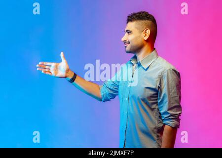 Ritratto di uomo felice in camicia stretching mano che accoglie gli ospiti con un sorriso toothy sul viso, Piacere di conoscerti. Studio interno girato isolato su colorato sfondo di luce al neon. Foto Stock