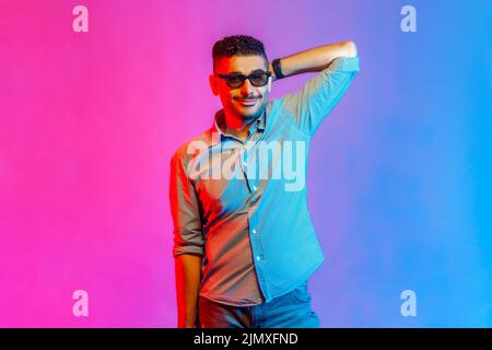 Ritratto di uomo in camicia e occhiali in piedi con mano rialzata e guardando la macchina fotografica con un sorriso positivo e espressione ottimista. Studio interno girato isolato su colorato sfondo di luce al neon. Foto Stock