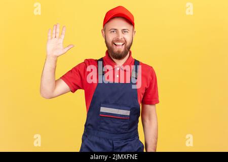 Ritratto di un uomo di lavoro felice in piedi e guardando la macchina fotografica, ondeggiante la mano, che dice ciao o Arrivederci, indossando tute blu e cappuccio rosso. Studio interno girato isolato su sfondo giallo. Foto Stock