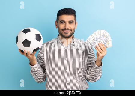 Sorridente uomo d'affari soddisfatto che tiene la sfera di calcio e ventilatore delle fatture del dollaro, scommesse sportive, grande vittoria, guardando la macchina fotografica, indossando la camicia a righe. Studio interno girato isolato su sfondo blu. Foto Stock