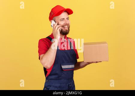 Ritratto di corriere positivo donna in piedi tenendo in piedi scatola di cartone, pacco per il cliente e parlare via telefono cellulare, indossare tute e tappo rosso. Studio interno girato isolato su sfondo giallo. Foto Stock