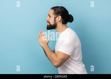 Per favore, sto pregando di perdonare. Vista laterale di un uomo sconvolto preoccupato con la barba che indossa una T-shirt bianca che cerca di implorare la disperata grimace, pregando per aiuto. Studio interno girato isolato su sfondo blu. Foto Stock