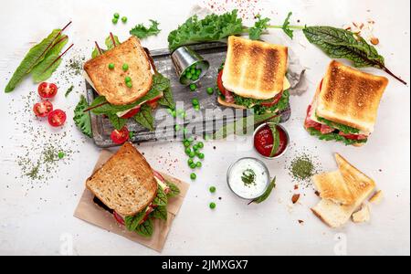Sandwich con formaggio, prosciutto, pomodoro e insalata fresca, fatto di pane integrale con crosta croccante su tavola di legno con salse su fondo bianco, Foto Stock