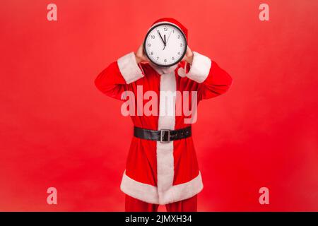 Ritratto di uomo sconosciuto in costume di babbo natale in piedi copertura viso con orologio a muro, sprecando il suo tempo, procrastinazione, organizzazione del tempo di lavoro. Studio interno girato isolato su sfondo rosso. Foto Stock