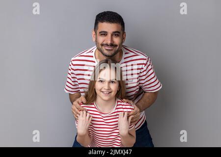 Ritratto di felice sorridente padre e figlia in T-shirt a righe in piedi e guardando la macchina fotografica, papà abbracciando il bambino piccolo, esprimendo la felicità. Studio interno girato isolato su sfondo grigio. Foto Stock