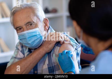 Professionista della salute femminile biracale che fa un'iniezione all'anziano caucasico a casa Foto Stock