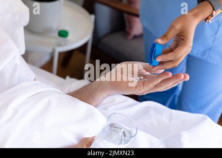 Sezione centrale dell'operatrice di salute femminile di biracial che dà le pillole mediche all'uomo anziano caucasico sul letto Foto Stock