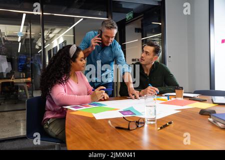 Un uomo d'affari caucasico che parla con gli smartphone mentre discute con i colleghi sui documenti Foto Stock