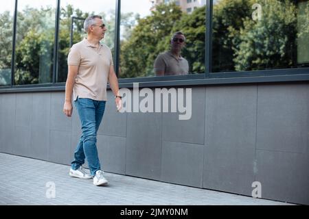 Vista laterale ritratto dal basso del vecchio uomo in occhiali da sole che camminano sulla strada lungo l'edificio degli uffici e guardando la silhouette riflesso nella finestra. Urbano Foto Stock