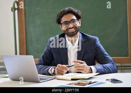 Buon giovane insegnante indiano di affari seduto al lavoro in classe. Verticale Foto Stock