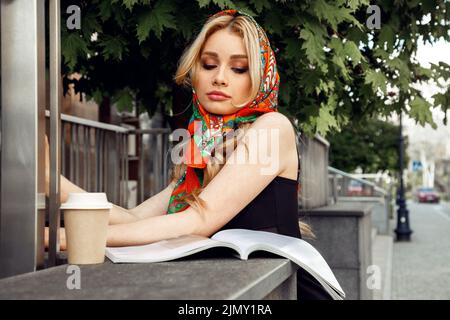 Ragazza romantica con trucco luminoso in elegante foulard colorato guardando verso il basso e rivista di lettura con un bicchiere di caffè in primo piano mattina. Stile parigino Foto Stock