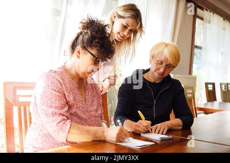 Giovane donna in piedi dietro due donne anziane, spiegando il business. Gli anziani sono seduti alla scrivania e annotano le note nel blocco note. Foto Stock