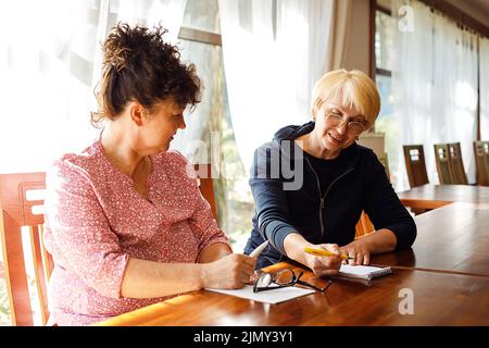 Ritratto di due donne anziane sorridenti sedute alla scrivania marrone, parlando, parlando, facendo appunti in una grande stanza. Foto Stock