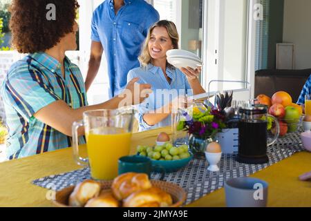 Giovane donna caucasica che passa piatti al giovane biraciale mentre fa colazione al tavolo da pranzo Foto Stock