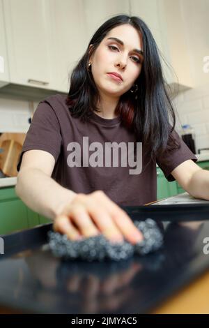 Giovane donna stanca con una spugna metallica per la pulizia, lava una teglia da forno in cucina, fuoco selettivo, verticale Foto Stock