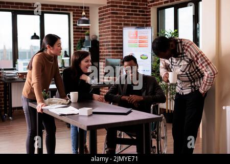 Imprenditore seduto in sedia a rotelle e parlare con i partner aziendali in pausa di lavoro, ridendo e divertirsi in un ufficio per disabili. Lavoratore di sesso maschile con disabilità che parla con i colleghi. Foto Stock