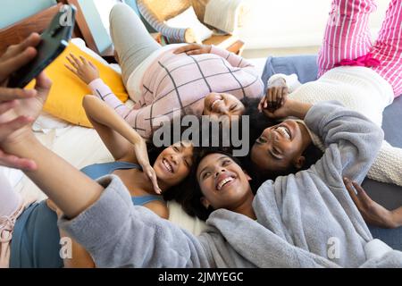 Vista ad angolo alto di giovane ragazza biraciale allegra che prende selfie con le amiche sdraiate a letto Foto Stock