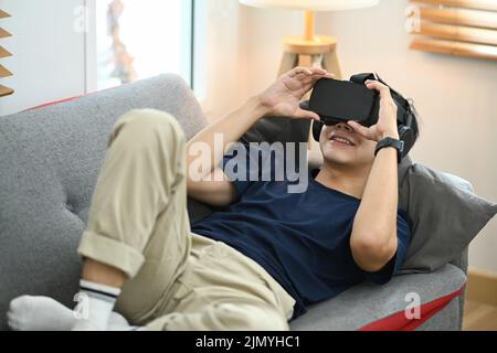 Uomo in abiti casual sdraiato sul divano, sperimentando opportunità di gioco con cuffie per realtà virtuale Foto Stock