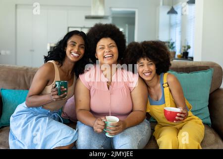 Ritratto di allegre giovani amici biraciali in possesso di tazze da caffè rilassante sul divano a casa Foto Stock