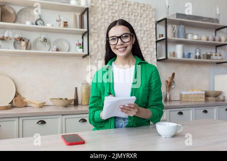 Psicologo online. Una giovane psicologa femminile specializzata in occhiali e una maglietta verde offre consulenze online ai pazienti tramite web camera. Guarda la fotocamera, parla, scrive in un notebook. Foto Stock