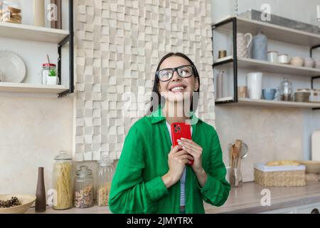 Giovane ragazza amorosa adolescente in occhiali e maglietta verde testando con la persona amata in rosso telefono a casa. Imbarazzata, lei nasconde il suo telefono, non lo mostra, sorride. Foto Stock