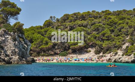 Spiaggia di baia sabbiosa a Minorca, Spagna, circondata da scogliere rocciose e pini, con acqua mediterranea turchese e bagnanti estivi Foto Stock