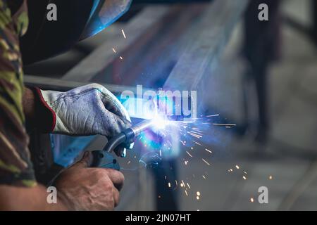 Un uomo che esegue la saldatura e la smerigliatura sul posto di lavoro in officina, mentre le scintille 'volano' tutto intorno a lui. Indossa un casco di protezione Foto Stock