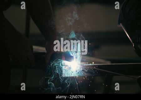 Un uomo che esegue la saldatura e la smerigliatura sul posto di lavoro in officina, mentre le scintille 'volano' tutto intorno a lui. Indossa un casco di protezione Foto Stock