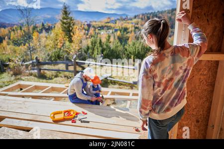 Padre, madre e figlio costruzione casa in legno cornice. Ragazzo toddler che aiuta il suo papà, mentre la donna che li cerca sul cantiere. Ragazzi che indossano casco e tute blu. Falegnameria, concetto di famiglia. Foto Stock