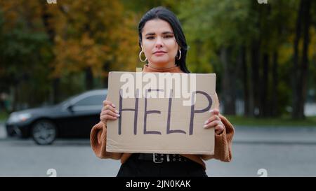 Primo piano triste giovane donna si alza all'aperto vicino strada tenendo segno di cartone con le parole AIUTARE sconvolto ragazza ispanica alla ricerca di lavoro esperienza Foto Stock