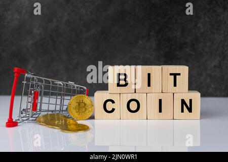 Bitcoin nel carrello dello shopping e blocchi di legno con la parola bitcoin su sfondo grigio scuro in tavola bianca. Concetto di valuta futura. Foto Stock
