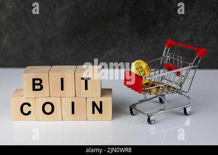 Bitcoin nel carrello dello shopping e blocchi di legno con la parola bitcoin su sfondo grigio scuro in tavola bianca. Concetto di valuta futura. Foto Stock
