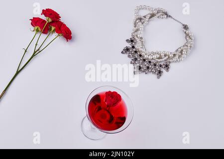 Accessori per donna a posa piatta su sfondo bianco. Cocktail rosso con fiori di rosa e collana. Foto Stock