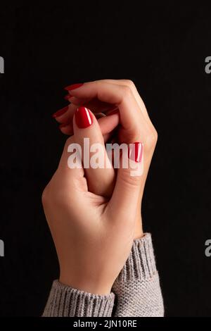 Maniche femminili in maglia grigia con manicure - chiodi rossi su sfondo nero scuro. Concetto di cura delle unghie Foto Stock