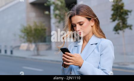Primo piano ritratto in città bionda seria ragazza business donna utente si trova sulla strada guardando il telefono cellulare dispositivo wireless gadget utilizzando l'app per smartphone Foto Stock