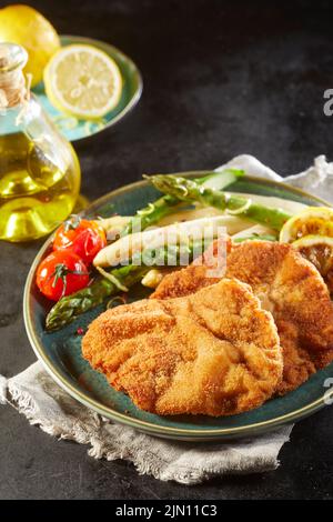 Da sopra schnitzel yummy con pomodori ciliegini al vapore e asparagi serviti su piatto vicino a bottiglia di olio e limoni freschi su sfondo nero Foto Stock